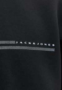 Tissu noir avec des lignes pointillées blanches au-dessus et en dessous du texte « JACK & JONES » imprimé en lettres blanches majuscules.