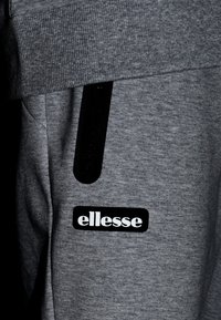 Pantalones de chándal grises hechos de un tejido suave, con un bolsillo con cremallera negra y un parche de logo negro que dice "ellesse" en el muslo.