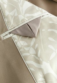 Tela beige y crema con un patrón floral, de textura suave y bordes cosidos en contraste. Incluye un detalle de bolsillo.