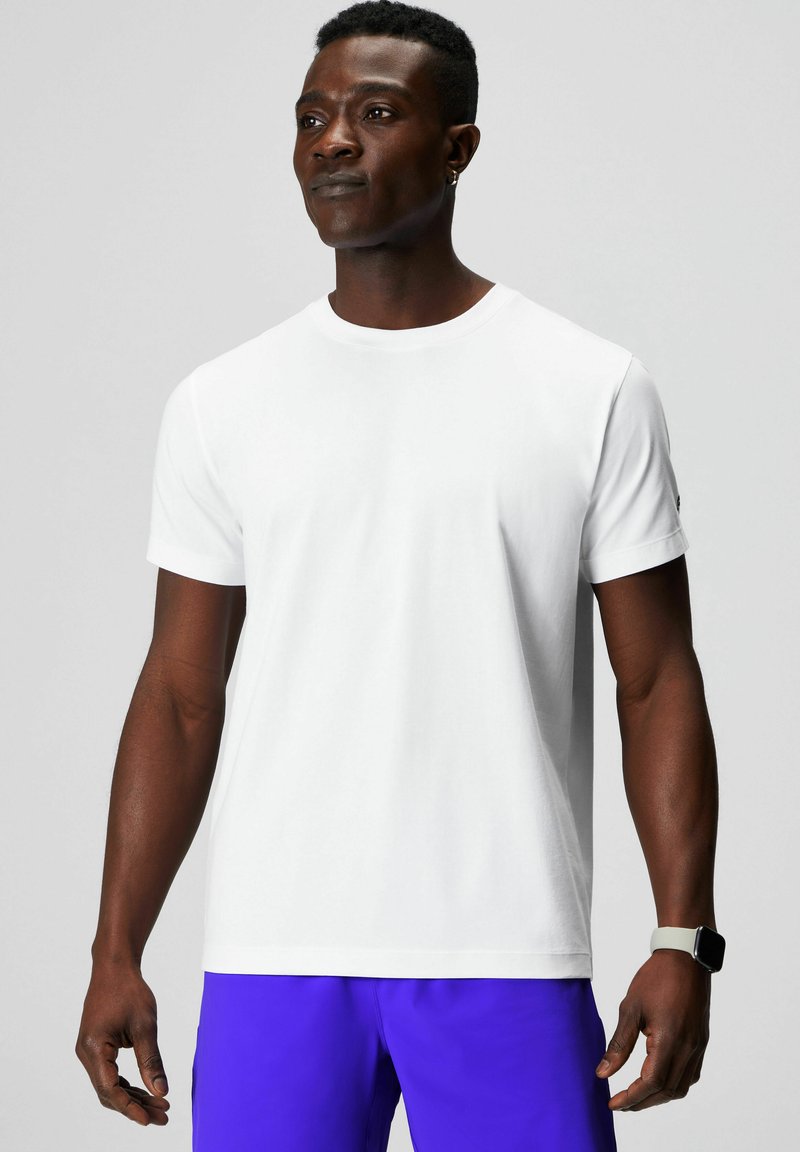 White short-sleeve t-shirt with a crew neck, made of smooth cotton fabric. Features a small logo on the left sleeve. Model wearing purple shorts.