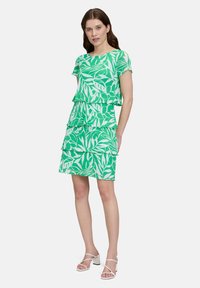 Grünes, gestuftes Kleid mit einem Blättermuster, kurzen Ärmeln und einem runden Halsausschnitt. Der Stoff hat eine glatte Textur, ergänzt durch weiße Blockabsatzsandalen.