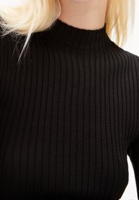 Personne blonde portant un haut noir côtelé à col montant, vue du menton jusqu'au haut du torse sur un fond uni.