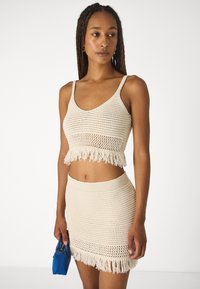 Beige strikket crop-top og skjørt med frynsete kant. Toppen har scoop-hals og justerbare stropper. Modellen holder en liten blå håndveske.