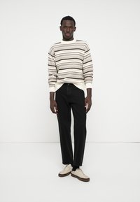 Homme portant un pull rayé crème et marron, un jeans noir et des baskets beiges, se tenant contre un fond blanc.