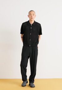 Black short-sleeve knit shirt with ribbed texture and buttons, paired with loose-fit black trousers and grey sneakers, against a plain background.