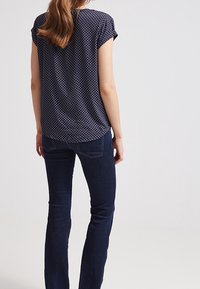 Tyrkysová košile s krátkým rukávem a geometrickým vzorem, spárovaná s tmavými úzkými džíny. Košile má volný střih a mírně zakřivený spodní lem.