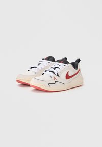 Sneakers Nike z beżową siateczką na górze, czarnymi akcentami, czerwonym swooshem, białymi sznurowadłami oraz teksturowaną gumową podeszwą w różowym odcieniu.