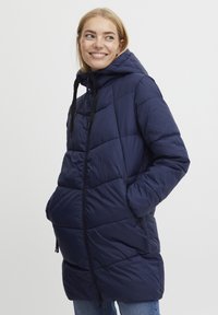 Marineblaue Steppjacke mit Kapuze, ausgestattet mit einem Reißverschluss, aufgesetzten Taschen und einem gesteppten Design. Glattes, leichtes Material.