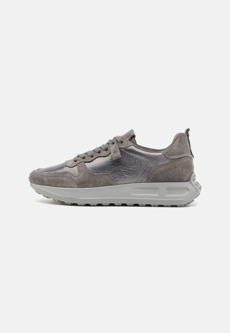 Zapatilla gris que presenta una combinación de materiales sintéticos suaves y ante texturizado, cierre de cordones y una suela de goma gruesa.