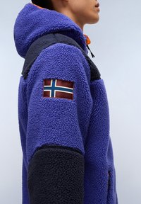 Polarowa kurtka w kolorach fioletowym i czarnym, z kapturem oraz naszywką z norweską flagą na rękawie. Strukturalny, ciepły materiał.