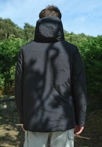 Must must jacket kõrge kaeluse ja kapuutsiga, sileda tekstuuri ja sobiva disainiga, nähtuna tagant roheluse taustal.