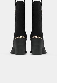 Botas de tobillo negras con una textura suave, acentos metálicos dorados en el talón y un diseño de tacón ligeramente acampanado. Patrones grabados en la parte superior.