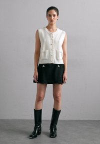 Balta adīta veste ar pogu aizdari un priekšējām kabatām, saskaņota ar melniem svārkiem, kurus rotā priekšējās pogas. Modelis nēsā melnas ceļa augstuma zābakus.