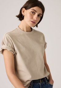 Γυναίκα με κοντά σκουρόχρωμα μαλλιά φοράει μπεζ μπλουζάκι με μανίκια που είναι διπλωμένα και μπλε τζιν με μαύρη ζώνη, ποζάροντας μπροστά σε απλό φωτεινό φόντο.