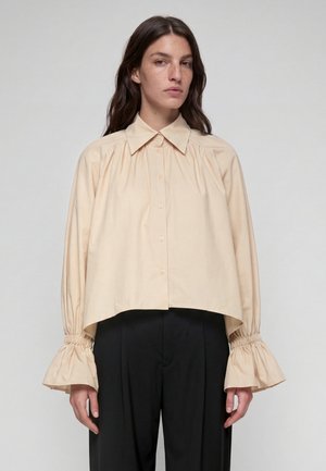 Frau trägt eine beige, knöpfbare Bluse mit gepufften, gerafften Manschetten und schwarze, hoch taillierte Hosen vor einem schlichten Hintergrund.