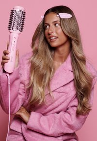 Brosse soufflante avec un manche en plastique rose et des poils noirs. Le modèle porte un peignoir en peluche rose, a de longs cheveux ondulés et des pinces à cheveux roses.