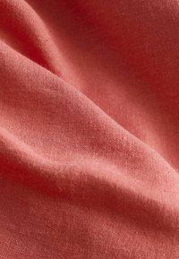 Tissu en lin corail avec une texture lisse, présentant de légères rides et des plis doux, soulignant sa légèreté et sa nature respirante.