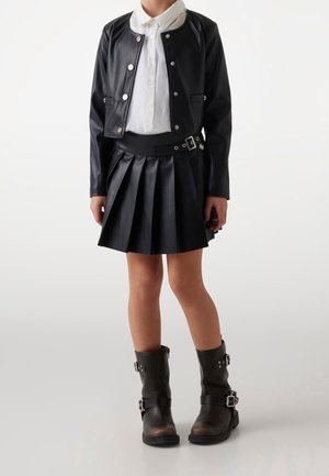 Barn iført sort kort læderjakke, hvid skjorte med knapper, sort lædernederdel med plisseringer og bælte samt sorte støvler med spænde.