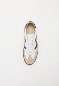 White athletic shoe with gray suede accents, black side stripes, and white laces. Tan rubber sole with a logo in gold on the insole.