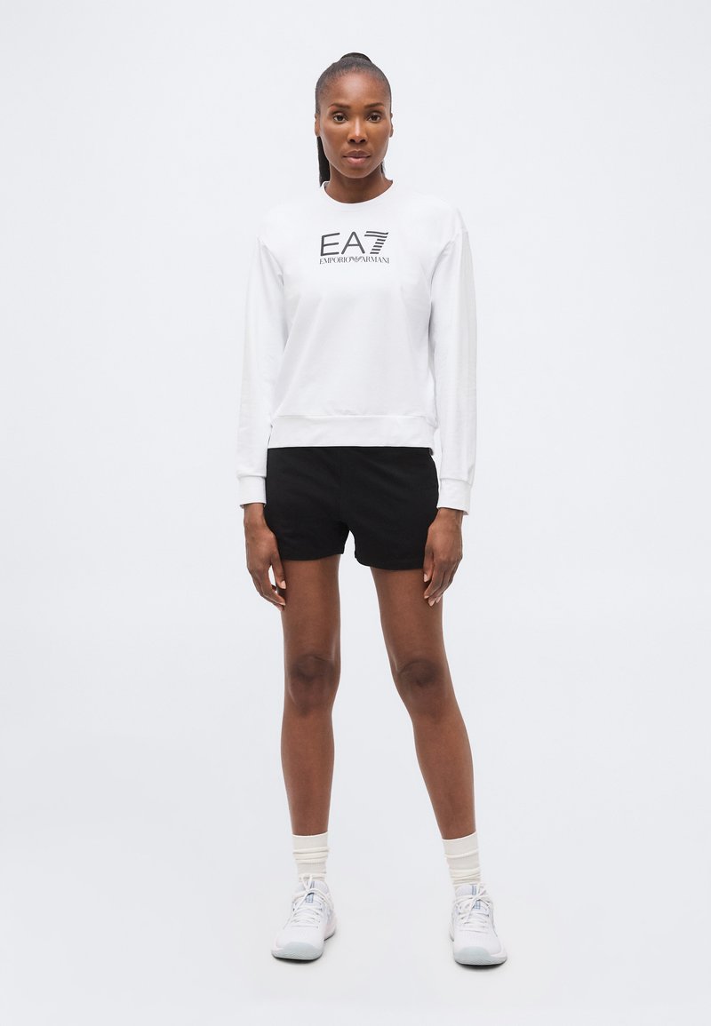 Γυναίκα στέκεται φορώντας λευκό φούτερ EA7 Emporio Armani, μαύρο σορτς, λευκές κάλτσες και λευκά αθλητικά παπούτσια σε απλό φωτεινό φόντο.