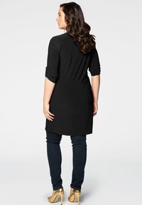 Sort lang tunika med opadvendte ærmer, parret med mørkeblå skinny jeans og guld højhælede sko. Glat tekstur, enkelt design og knælængde.