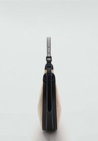 Sac en toile beige avec accents en cuir noir, doté d'un profil mince et d'une sangle ajustable. Remarquable pour son design minimaliste et ses variations de texture.