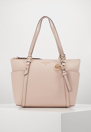 Sac cabas en cuir beige avec poignées doubles, poches latérales, logo Michael Kors doré et breloque pompon en cuir sur une surface blanche.