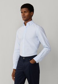 Uomo che indossa una camicia azzurro chiaro abbottonata, infilata in pantaloni blu scuro, in piedi con una mano in tasca contro uno sfondo semplice.