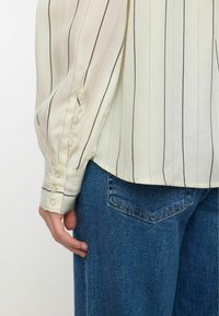 Knoopshirt in crème met zwarte strepen; heeft opgezette mouwen en vijf knopen bij de manchetten. Gecombineerd met blauwe denimstof.