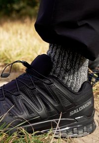 Black hiking shoe with textured upper, lace system, and padded collar, paired with a gray speckled sock, set against grassy terrain.