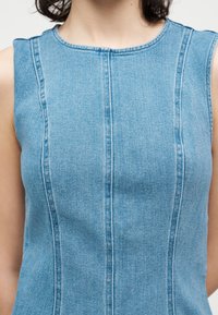 Αμάνικο μπλε τζιν τοπ με κάθετες ραφές που φοράει άτομο με σκούρα σγουρά μαλλιά πάνω σε λευκό φόντο.