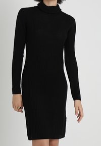 Robe en maille côtelée noire avec manches longues et col roulé. La robe est ajustée et présente des fentes latérales près de l'ourlet.