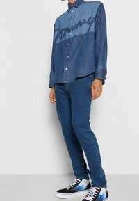 Denim button-up shirt with a blue gradient, featuring a large logo print, paired with dark blue jeans and black sneakers with blue accents.