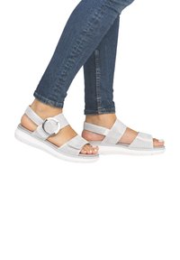 White strappy sandals with woven texture, silver hardware, and cushioned sole, paired with fitted blue jeans.