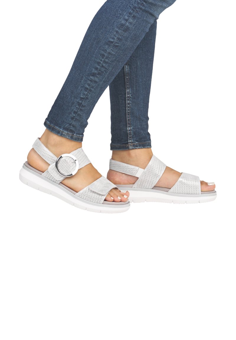 White strappy sandals with woven texture, silver hardware, and cushioned sole, paired with fitted blue jeans.