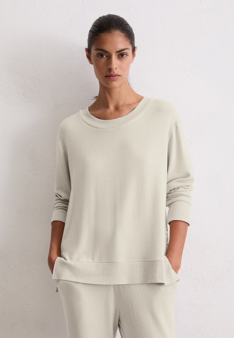 Femme aux cheveux bruns tirés en arrière, portant un pull crème surdimensionné et un pantalon assorti, se tenant devant un mur blanc texturé uni.