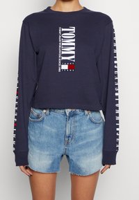 Námořnická modrá krátká mikina s dlouhým rukávem a bílým textem a logem. Kombinováno se světlými modrými džínovými šortkami, které ukazují ležérní design.