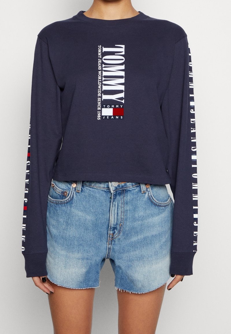 Námořnická modrá krátká mikina s dlouhým rukávem a bílým textem a logem. Kombinováno se světlými modrými džínovými šortkami, které ukazují ležérní design.