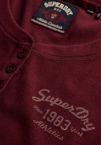 Maroon ribbed fabric shirt with buttons, printed "SuperDry 1963" and label reading "SuperDry NYC Athletic Essentials."