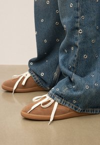Jeans med silverfärgade öglor och vida ben, kombinerat med bruna mockaskor med vita snören och gummisålar.