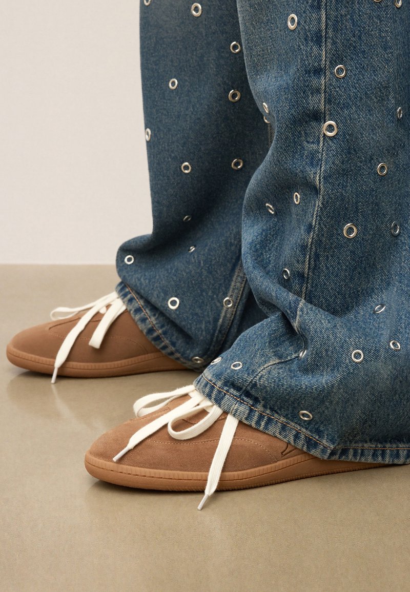 Jeans med silverfärgade öglor och vida ben, kombinerat med bruna mockaskor med vita snören och gummisålar.