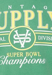 Zelené bavlněné tričko s bílým a žlutým textem, obsahuje "Vintage Supply", "Super Bowl Champions" a emblém s propletenými písmeny.