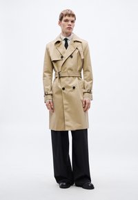 Béžový trenčkot s dvojradovým zapínaním, opaskom v páse a ramennými epolety. Nosený cez čierne široké nohavice a topánky.