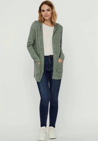 Groene gebreide cardigan met een capuchon, lange mouwen en twee voorkruiken, gedragen over een witte top en gecombineerd met donkere jeans en witte sneakers.