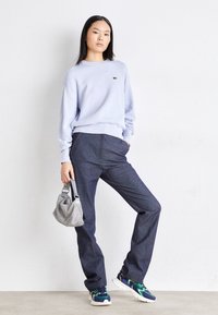 Light purple sweatshirt, navy trousers, and patterned sneakers. Model holds a small silver handbag. Simple, modern design with relaxed fit.