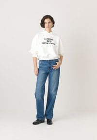 Biała bluza z kapturem z czarnym napisem, w połączeniu z niebieskimi prostymi dżinsami. Tkanina wydaje się być miękka i luźna. Czorne buty dopełniają look.