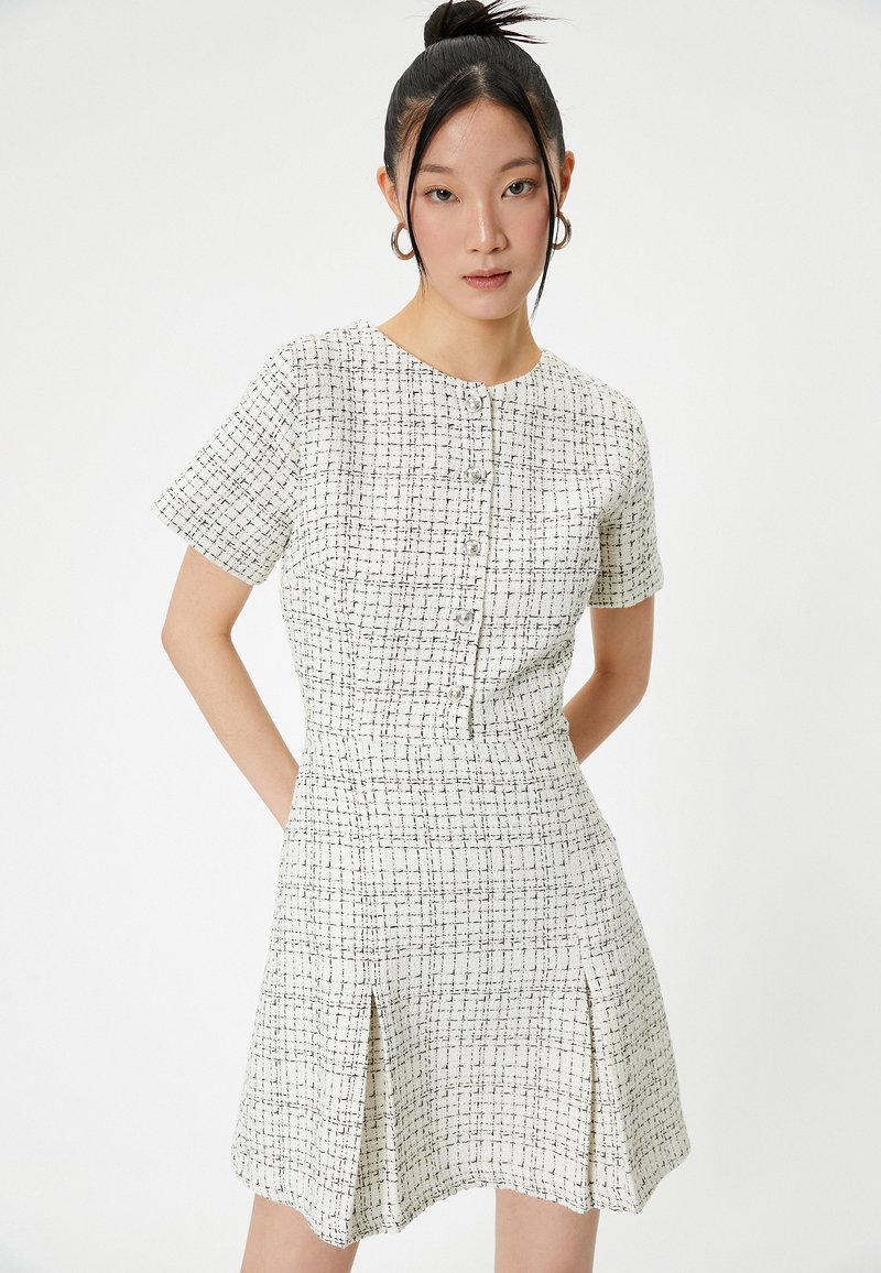 Vestido blanco con patrón de cuadros negros, mangas cortas, escote redondo, cierre frontal con botones y pliegues laterales para un detalle añadido.