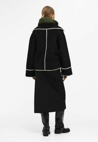 Giacca nera con texture e bordi bianchi, accoppiata a una gonna nera unita. La modella indossa stivali neri lucidi fino al ginocchio e una sciarpa verde.