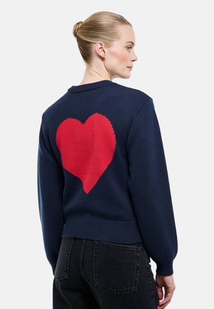 Femme aux cheveux blonds attachés en chignon portant un pull bleu marine avec un grand cœur rouge dans le dos, associé à un jean noir.