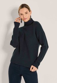 Frau in einem dunklen gerippten Rollkragenpullover und dunklen Hosen, die mit einer Hand nahe ihrem Gesicht und der anderen auf ihrer Hüfte posiert.